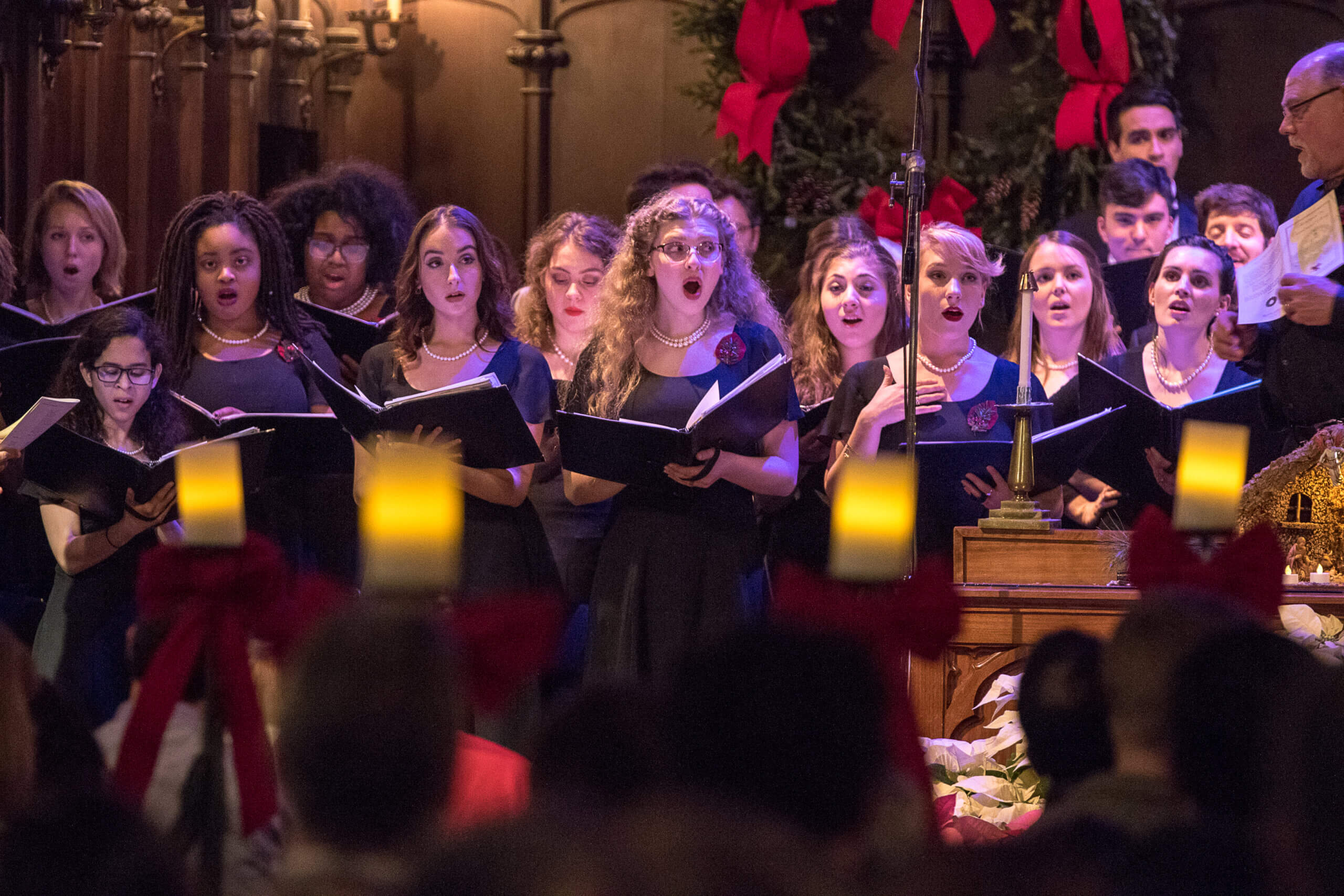 Young women in black dresses sing in a choir with candles in the foreground.
