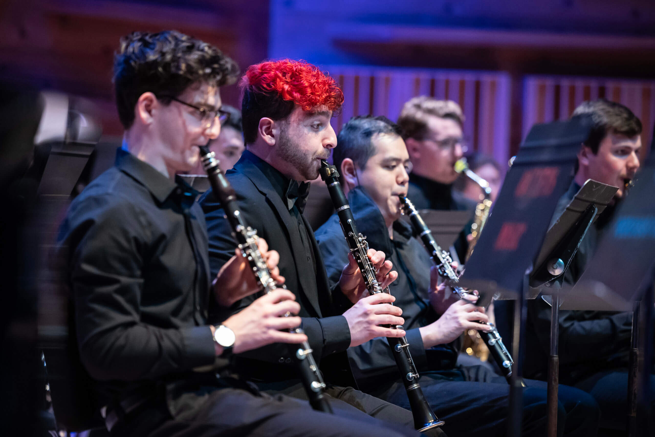 Three students play a wind instrument on stage in the foreground.The student in the middle has vibrant red hair.