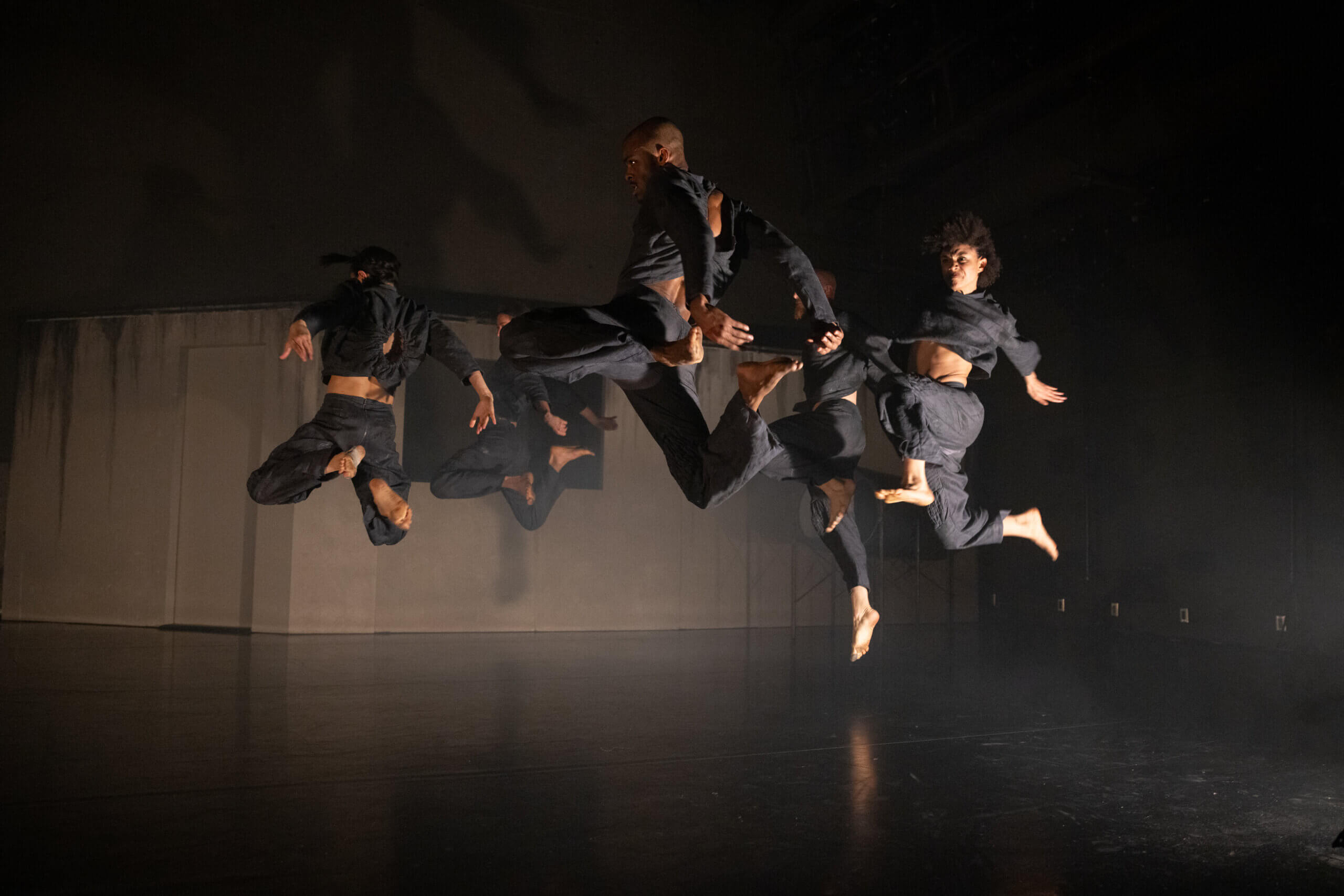 Dancers jump in a circle, spotlighted on stage. They are dressed in all-black.