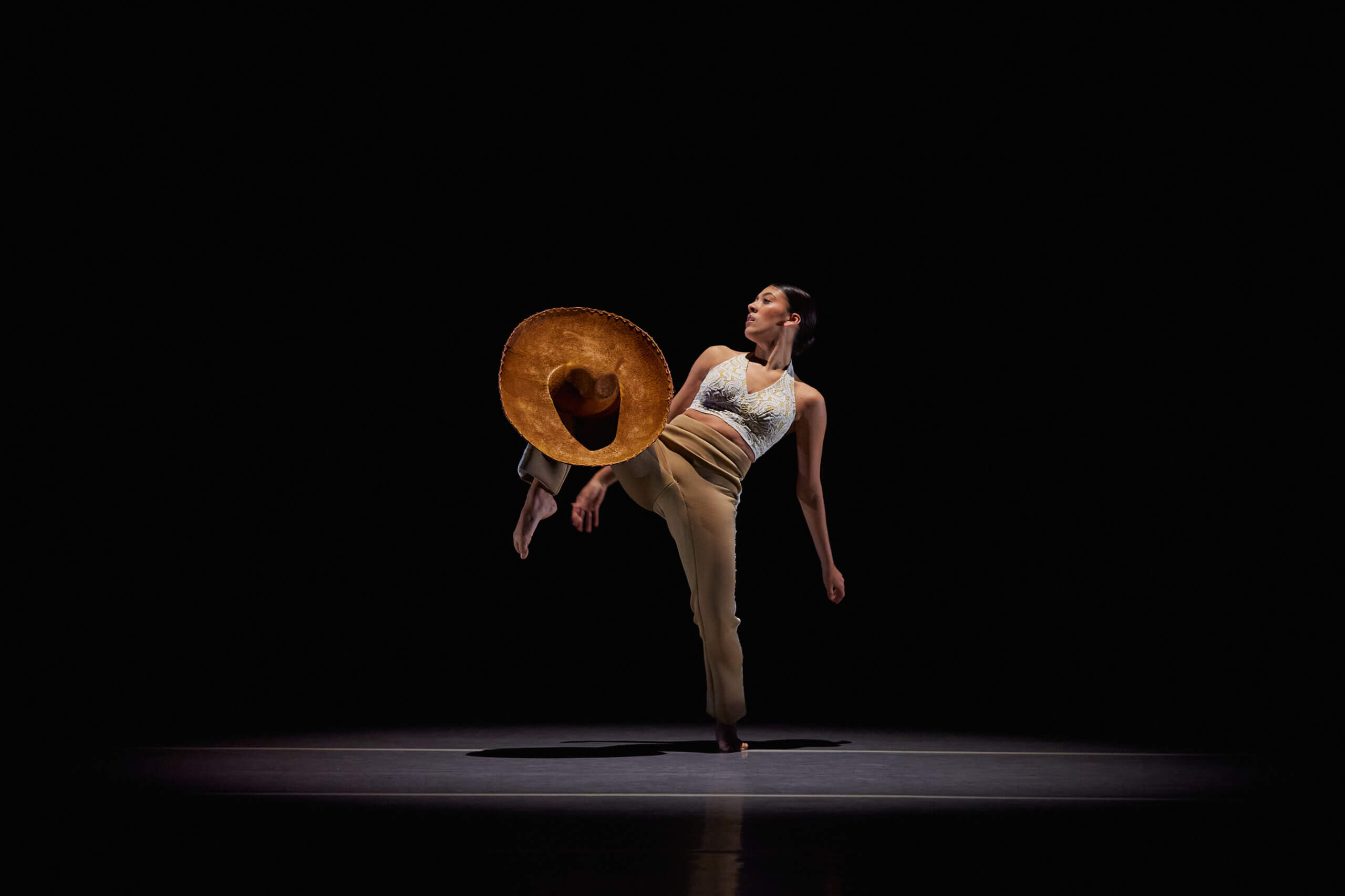 A dancer performs onstage under a spotlight. She's wearing a white top and brown pants, and is balancing a sombrero on her knee.