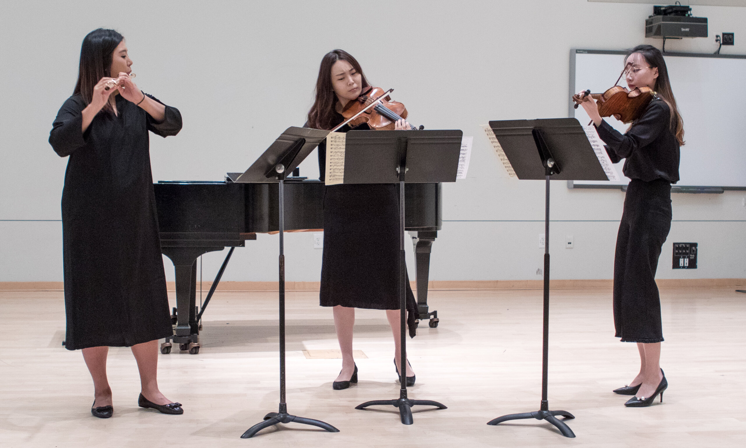 Three women perform instruments in front of music stands. 