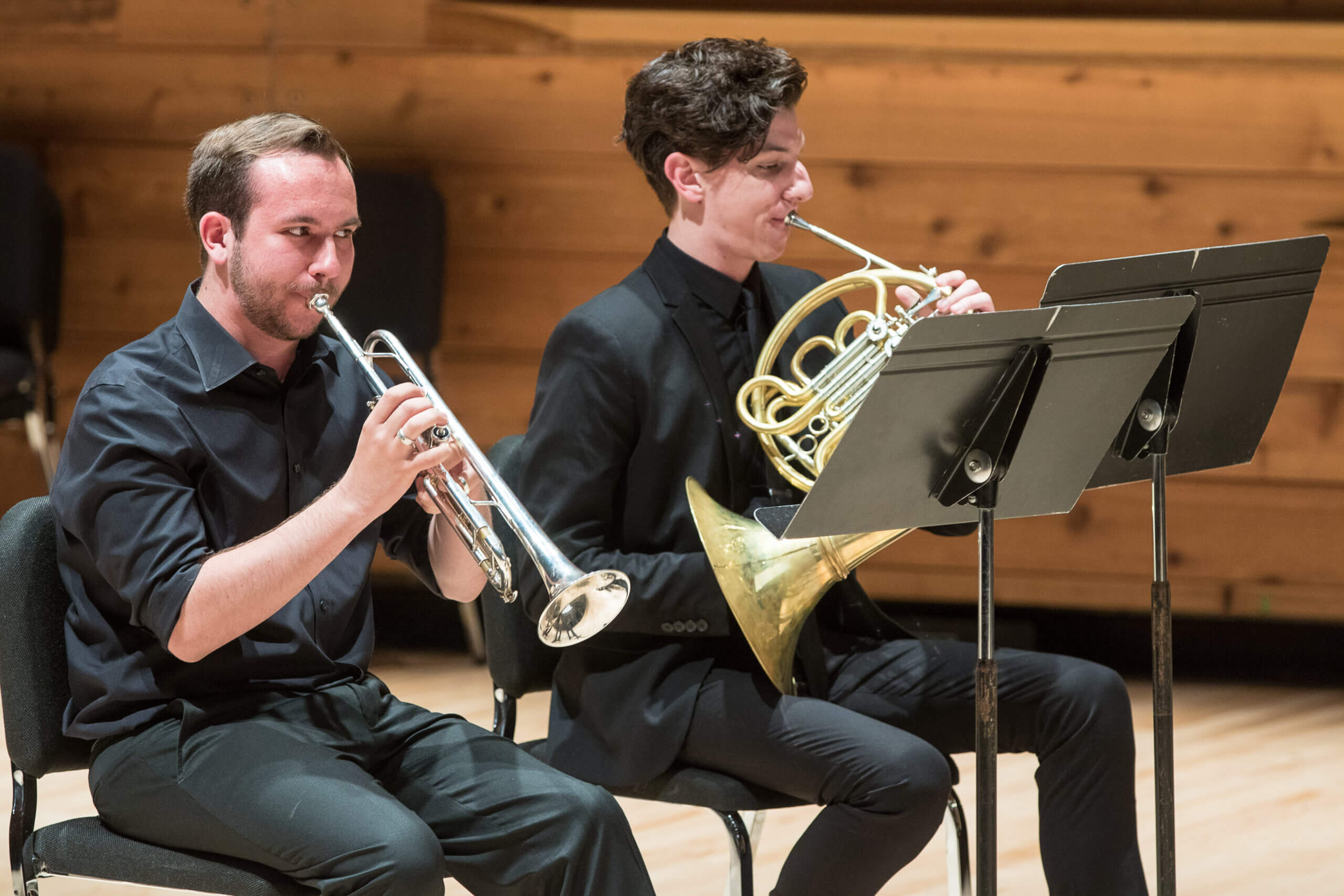 Two students perform on stage wearing all black. One plays the trumpet, and the other plays the French horn. 